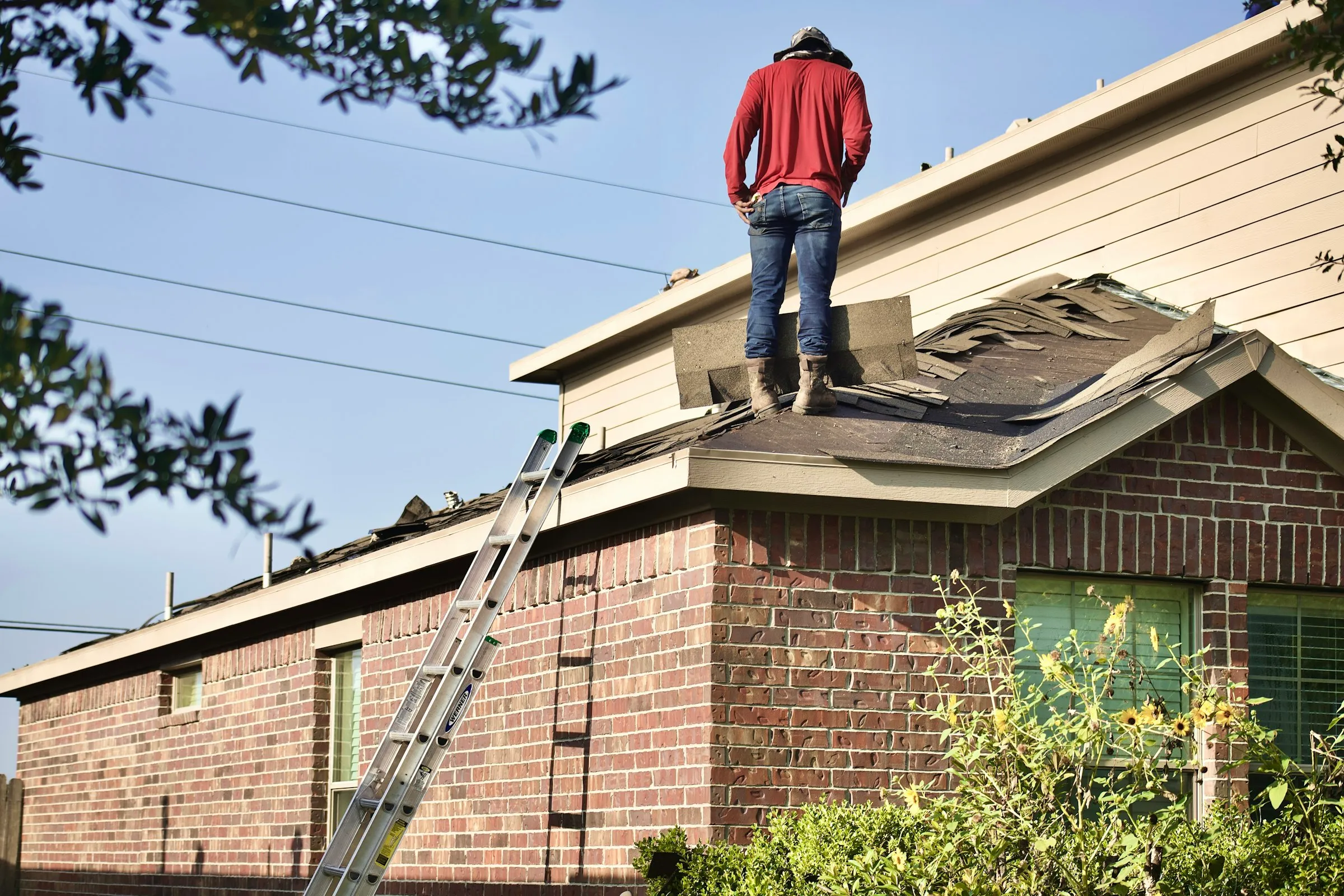 A roofer in a red shirt standing on a half stripped roof, with a ladder up against the brick wall of the house